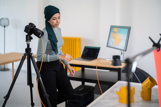 A woman in a headscarf works in a photo studio, reviewing images on a monitor and laptop. She is preparing for a product shoot featuring yellow objects as part of her photography work.