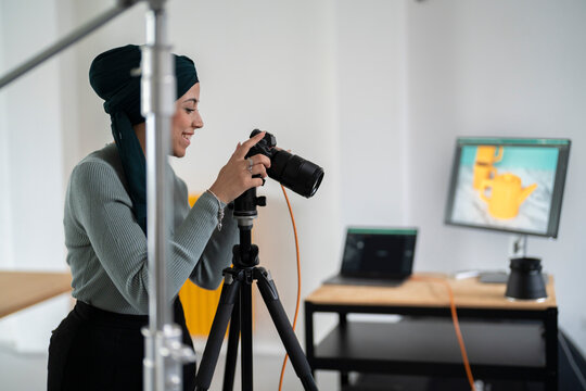 A woman in a headscarf is taking a product photo with a camera on a tripod in a studio setting. She is likely capturing images for e-commerce or marketing purposes, as seen on the monitor display.