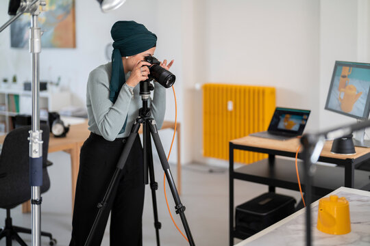 A woman in a headscarf photographs a product using a camera on a tripod in a studio. She reviews the images on monitors, likely preparing them for online sale or promotion.