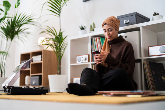 A woman in a headscarf sits on the floor, engrossed in her phone. She is surrounded by plants, a record player, and vinyl records, creating a cozy atmosphere for relaxation and digital connection.
