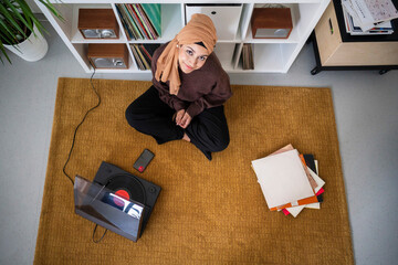 A woman in a headscarf sits on a rug, enjoying music from her turntable. She is surrounded by records, relaxing and listening to her favorite tunes at home.