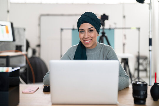A smiling woman in a hijab sits at her laptop in a photo studio. She is likely managing her photography business or editing photos, looking happy and engaged in her work.