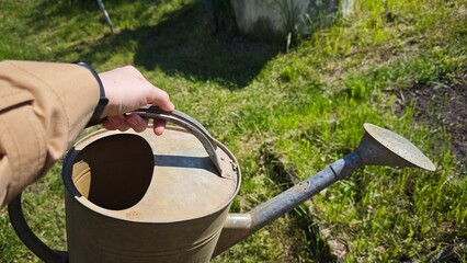 Hand holding a vintage metal watering can over green grass in a sunny garden. Concept of gardening routine and sustainable living