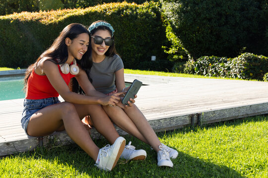 Smiling diverse female friends leaning over smartphone on pool deck, with headphones and sunglasses