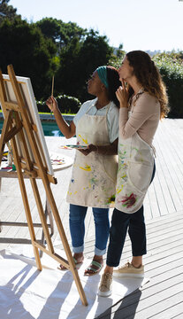 Painting Diverse female instructor and student holding brush and palette at pool deck, with easel