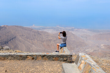  Woman sitting on a stone barrier taking a photo of breathtaking mountain and sea view from Jebel Jais, the highest peak in the United Arab Emirates