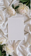 Fototapeta premium Blank greeting card surrounded by white peonies on silky fabric 
