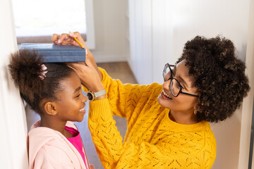 Diverse mother and daughter measuring height on door frame in hallway, with book and pencil