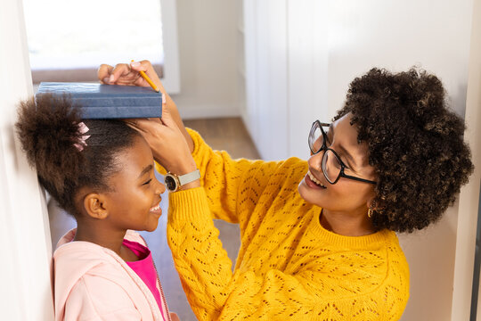 Diverse mother and daughter measuring height on door frame in hallway, with book and pencil