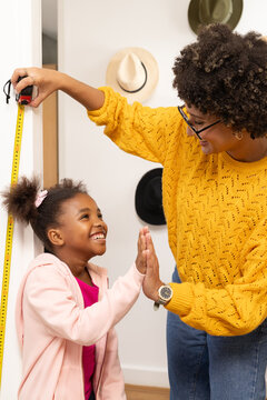 Diverse mother and daughter measuring height high fiving in hallway with tape measure, copy space