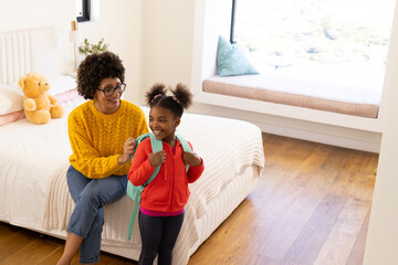 Fastening straps Diverse mother and daughter sitting in bedroom adjusting teal backpack