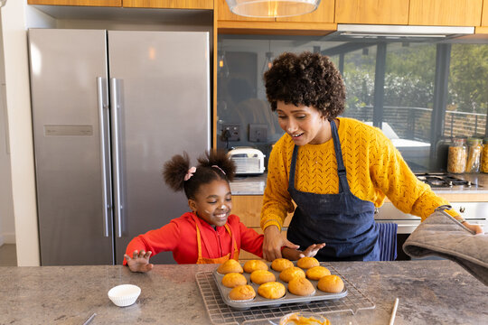 Standing Diverse mother and daughter smiling at kitchen island, with cooling rack of baked muffins