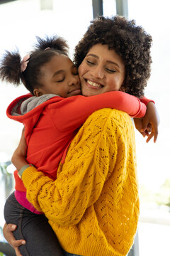 Hugging Diverse mother and daughter indoors near window, with yellow sweater and red hoodie