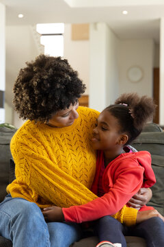 Hugging diverse mother and daughter sitting on dark gray sofa in living room, with cushions