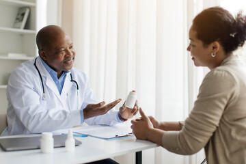 Fototapeta premium Smiling Black Doctor Man Demonstrating Prescribing Medications To Female Patient After Check Up In Clinic, Friendly Male Therapist Holding Bottle With Pills Or Vitamins And Showing It To Young Woman
