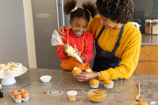 Baking mother and daughter piping icing onto cupcakes at kitchen island, with rainbow sprinkles