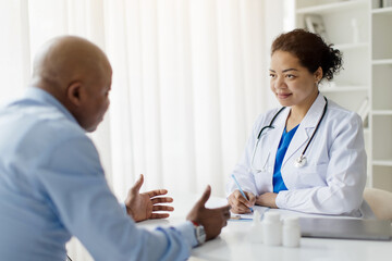 Smiling Black Doctor Woman In Uniform Consulting Male Patient During Meeting In Clinic, Female Therapist Wearing White Coat And Stethoscope Sitting At Desk And Listening Mature Man
