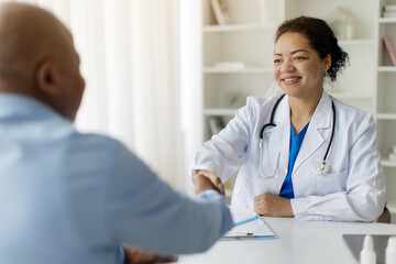 Fototapeta premium Medical Support Concept. Doctor Woman And Black Male Patient Handshaking During Meeting, Smiling Female Therapist In White Coat Welcoming Mature African American Man In Her Office, Closeup