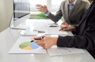 Cropped shot of two female colleagues analyzing financial data and discussing project results at desk with charts and graphs. Company employees engaged in teamwork with documents together in office.