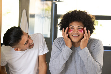 Applying pink under-eye patches, Diverse male friends laughing inside bathroom, with large window