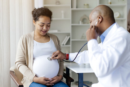 Male Gynecologist With Stethoscope Examining Belly Of Black Pregnant Female Patient, Obstetrician Doctor Listening Baby's Heartbeat While Making Check Up To Pregnant Woman In Maternity Clinic