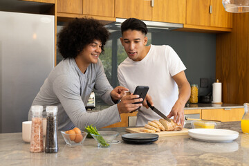 Diverse male friends preparing breakfast in modern kitchen, using smartphone and kitchen knife