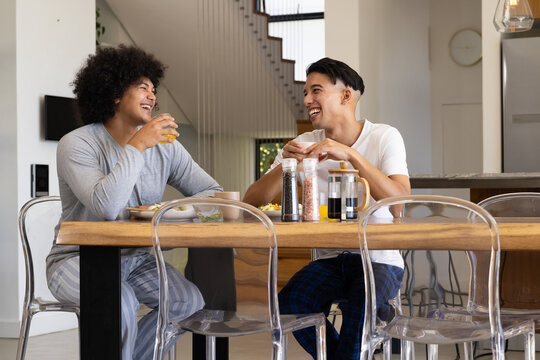 Laughing Diverse male friends sitting at wooden dining table, with orange juice, breakfast plates - Powered by Adobe