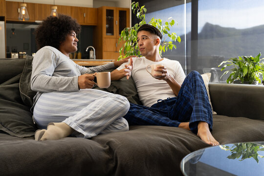 Chatting diverse male friends relaxing on sectional sofa in open-plan living room, with coffee mugs - Powered by Adobe