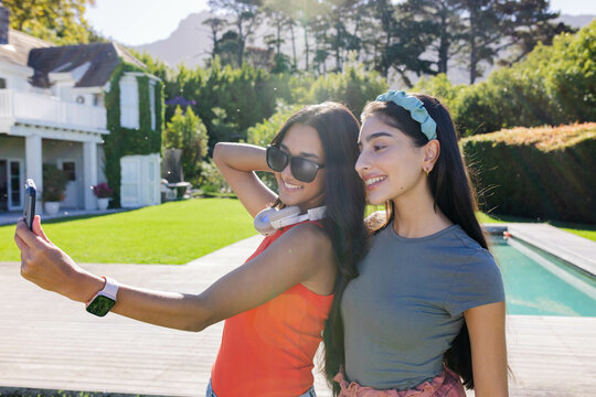 Taking selfie Diverse female friends posing on wooden deck by pool, with smartphone and smartwatch