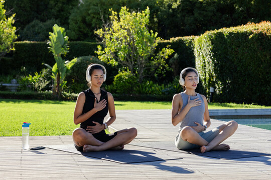 Meditating diverse female friends sitting on wooden deck by pool, with water bottle, smartphone