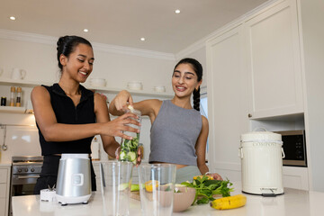 Diverse female friends adding cucumber and greens into blender in home kitchen, with compost bin