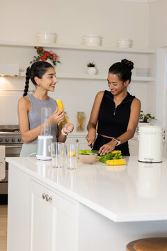 Preparing diverse female friends peeling banana and chopping greens in kitchen, with blender