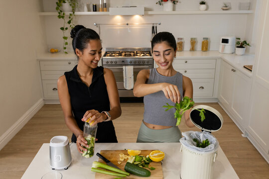 Diverse female friends mixing smoothies and filling compost bin at kitchen island, with blender jar - Powered by Adobe