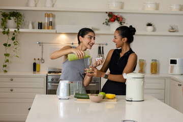 Diverse female friends pouring green smoothie into blender on kitchen island, with cucumber, lemon