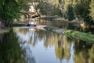 Obraz premium A serene river scene featuring a small stone dam with water gently cascading over the edge, surrounded by greenery and reflected sunlight, creating a peaceful and natural atmosphere. Serta, Portugal.