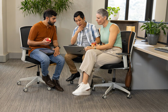Sitting Diverse coworkers holding red apples and using laptop in office break area, with tablet
