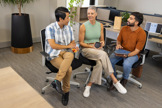 Diverse coworkers chatting in modern office meeting area, with jar of fruit and tablet