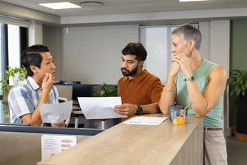 Discussing printed charts and data sheets at reception desk, diverse colleagues snacking on fruit