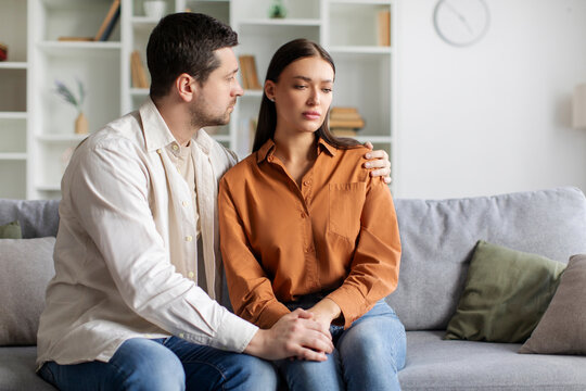 Loving man comforting his upset wife, husband providing support and expressing empathy while sitting together on couch in living room