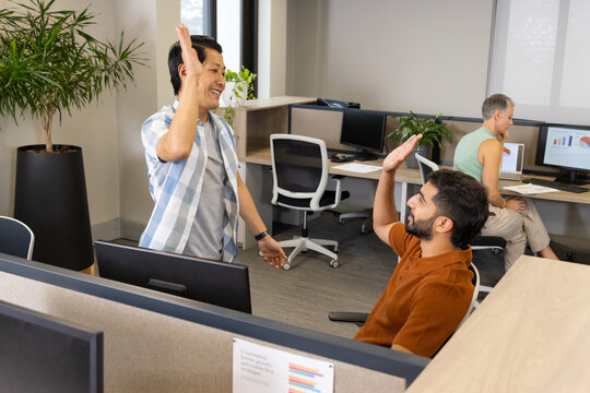 High-fiving Asian male coworkers working at open-plan office, with dual monitors and potted plant
