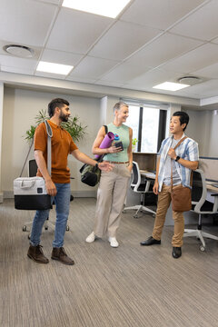 Chatting Diverse coworkers standing in modern open-plan office, with yoga mat and shoulder bags