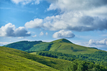 A lush green mountain range stretches under a dynamic sky filled with billowing clouds. Rolling hills, forested slopes, and a distant peak create a tranquil and scenic landscape summer Carpathian.