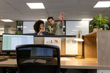 Diverse coworkers discussing spreadsheet data on monitor behind cubicle wall at office, with plants