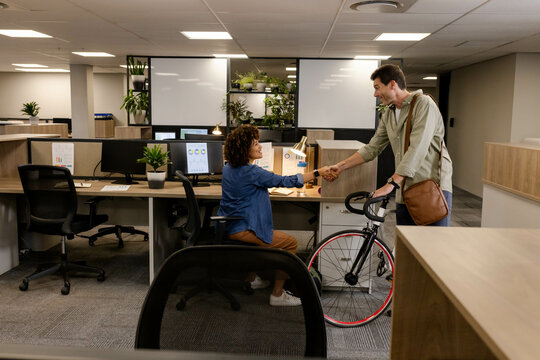 Diverse coworkers shaking hands at open-plan office workstation, with bicycle and dual monitors - Powered by Adobe