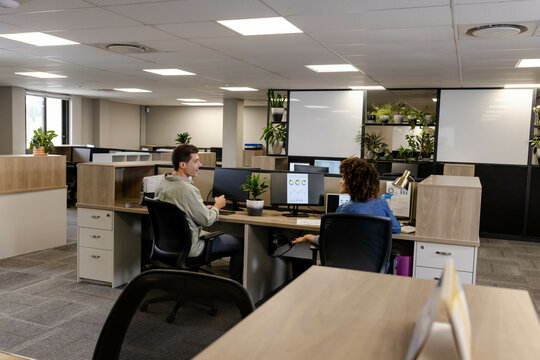 Collaborating diverse coworkers discussing ideas at open-plan office, with dual monitors and plants