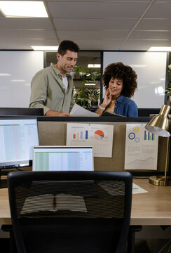 Diverse coworkers discussing documents in office cubicles, with monitors and charts