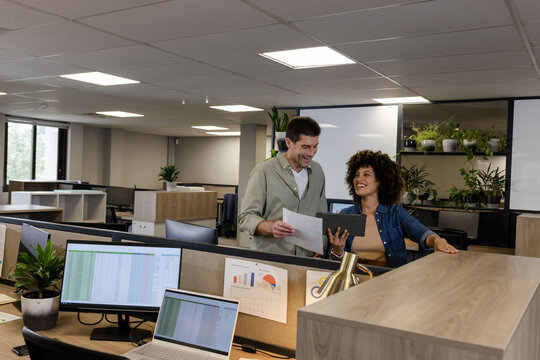 Diverse coworkers reviewing charts at tall desk in office, with tablet, dual monitors and laptop