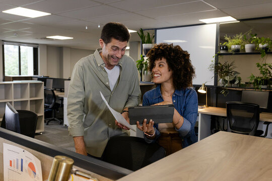 Diverse coworkers standing and reviewing documents in open-plan office, with tablet and paper
