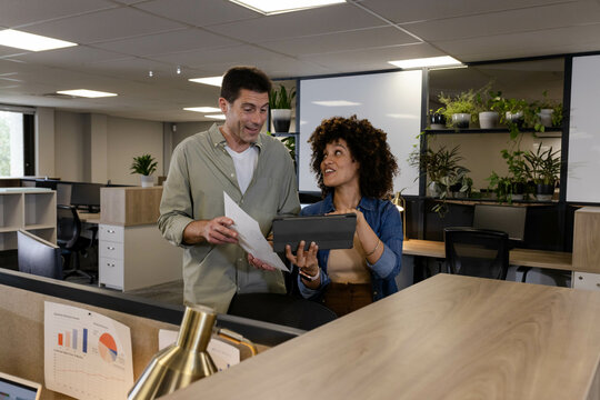 Diverse coworkers collaborating over printed papers and tablet at workstation, with potted plant