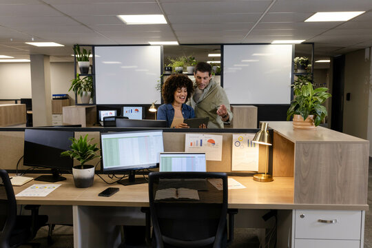 Diverse coworkers reviewing spreadsheet data on tablet in office cubicle, with monitors and charts - Powered by Adobe